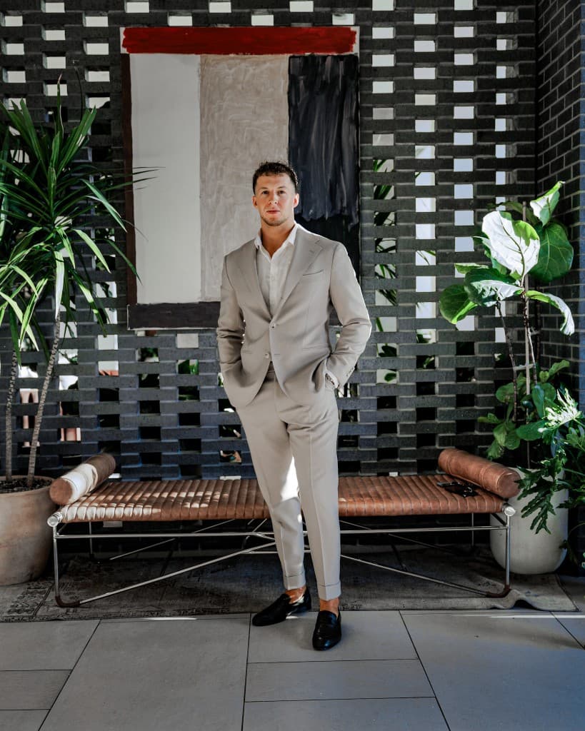 Hayden Mitchell, founder of RevCore HQ. Editorial portrait in a tailored light beige suit over an open-collar white shirt and black loafers, hands in pockets, standing in front of a red-white-black abstract painting on a perforated black brick wall flanked by tropical plants.